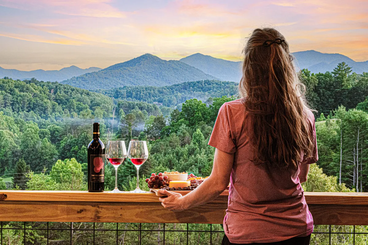 A beautifully arranged food platter and two wine glasses rest on a table overlooking expansive mountain vistas, bathed in warm sunset light. The setting suggests an intimate wine tasting or gourmet picnic experience with the mountains as a breathtaking backdrop.