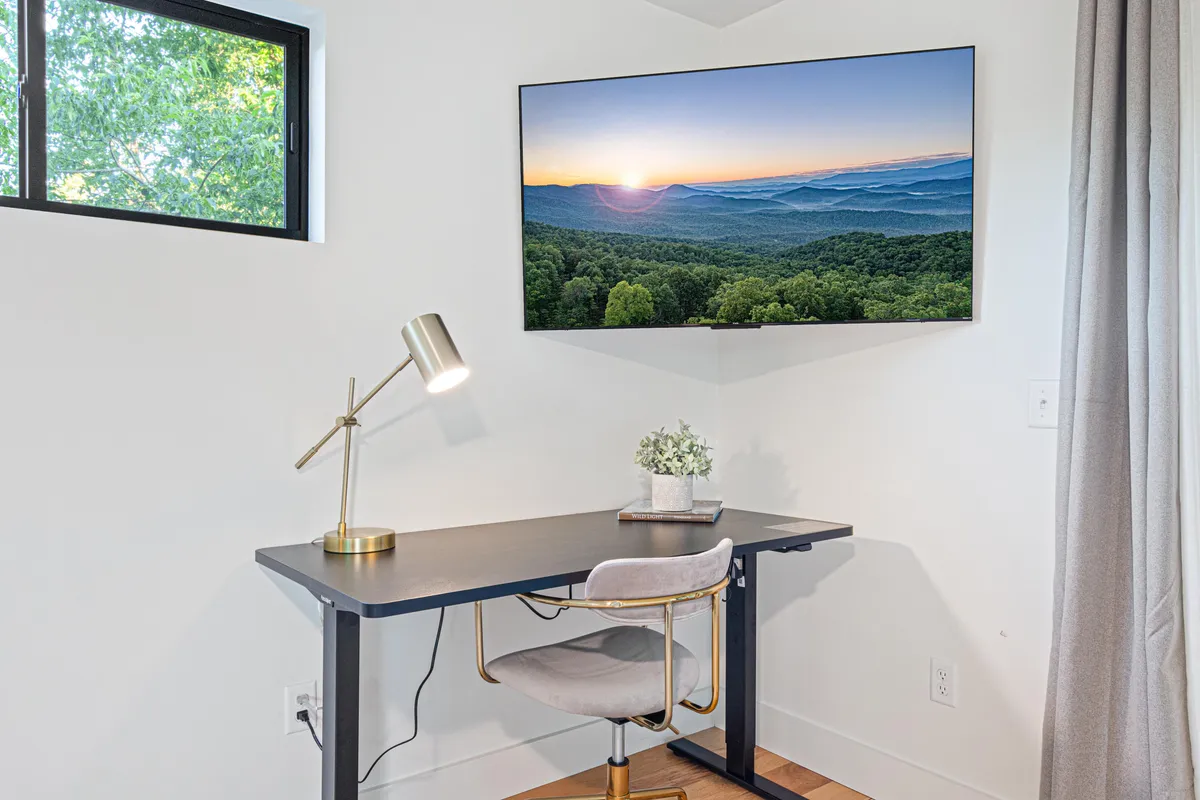 A serene workspace featuring a desk, television, lamp, and window that fills the space with natural light. The window offers a scenic backdrop, creating an inviting area for focused work or creative pursuits.