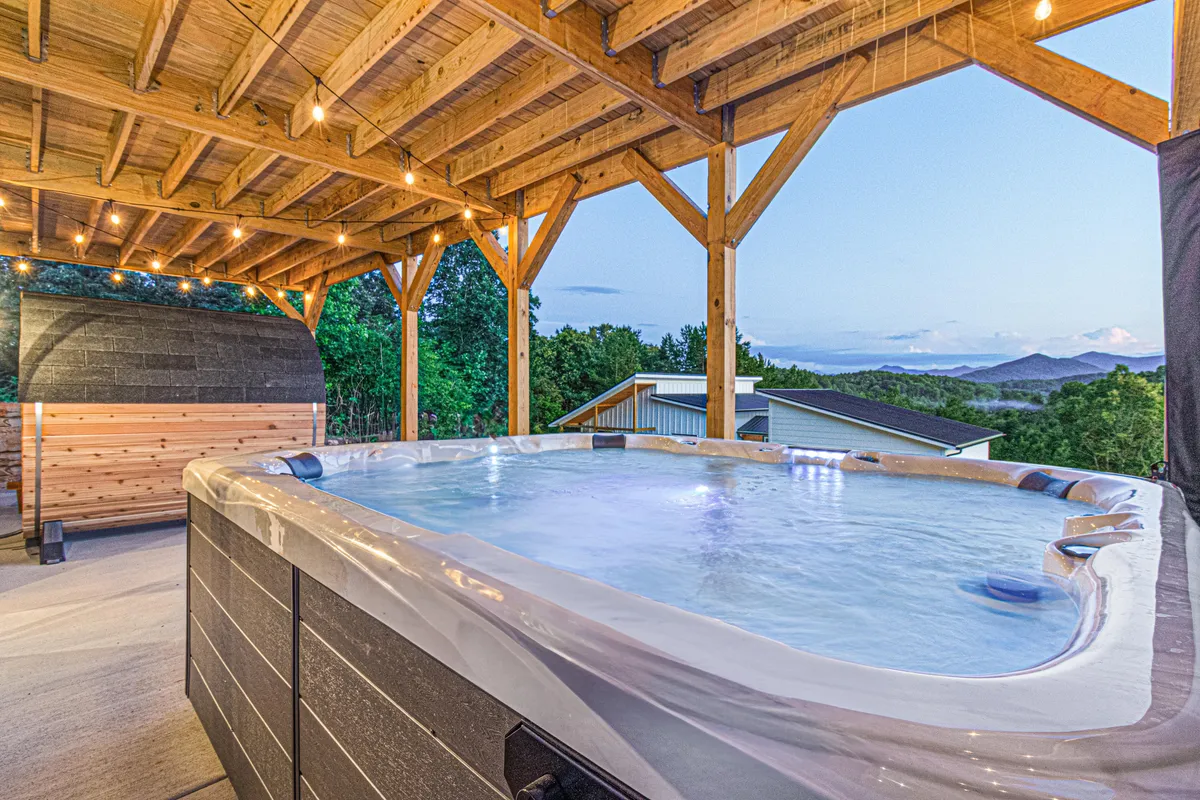 An inviting outdoor hot tub sits atop a deck framed by wooden beams and soft, ambient lighting, all set against a backdrop of sweeping mountain views. The scene is captured at dusk, capturing the tranquil transition from day to evening in the Blue Ridge Mountains.