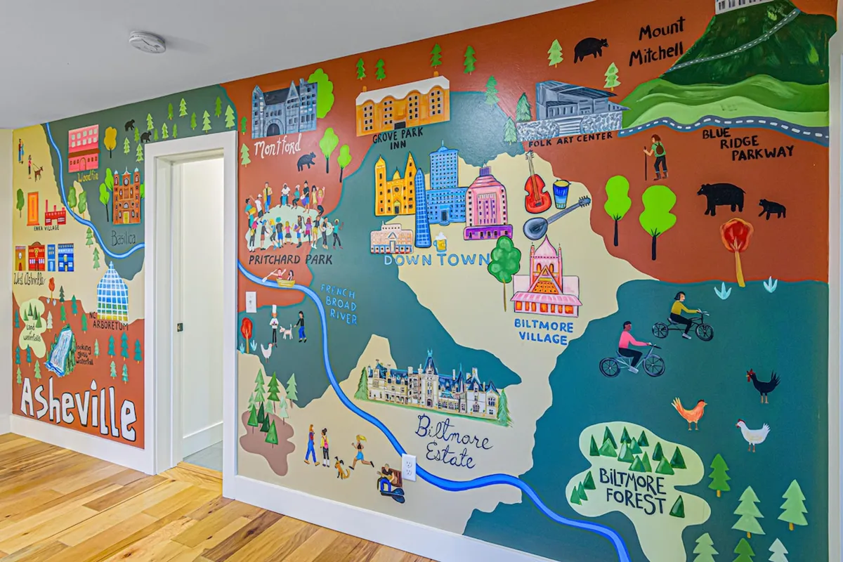 A vibrant, playful interior mural features a map of Asheville and illustrated highlights of the area, including nods to art and culture. The scene encourages discovery of the local arts, food, and attractions, set within a welcoming, creative space.