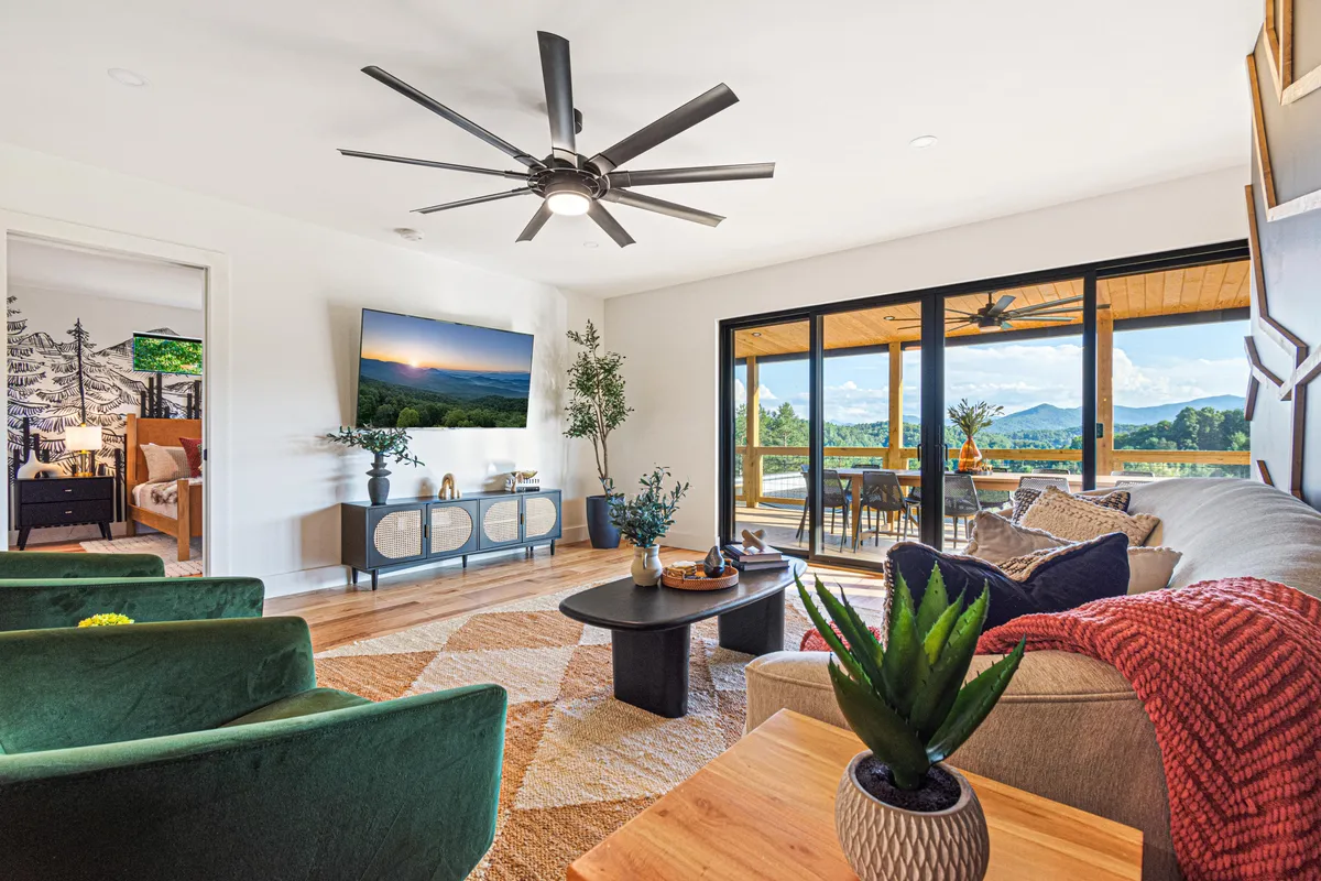 A sophisticated living room with expansive floor-to-ceiling windows that offer breathtaking views of the Blue Ridge Mountains. The space features modern green velvet chairs, a stylish coffee table, plush seating, decorative plants, and a large TV. Natural light floods the room, highlighting high-end finishes and tasteful decor.