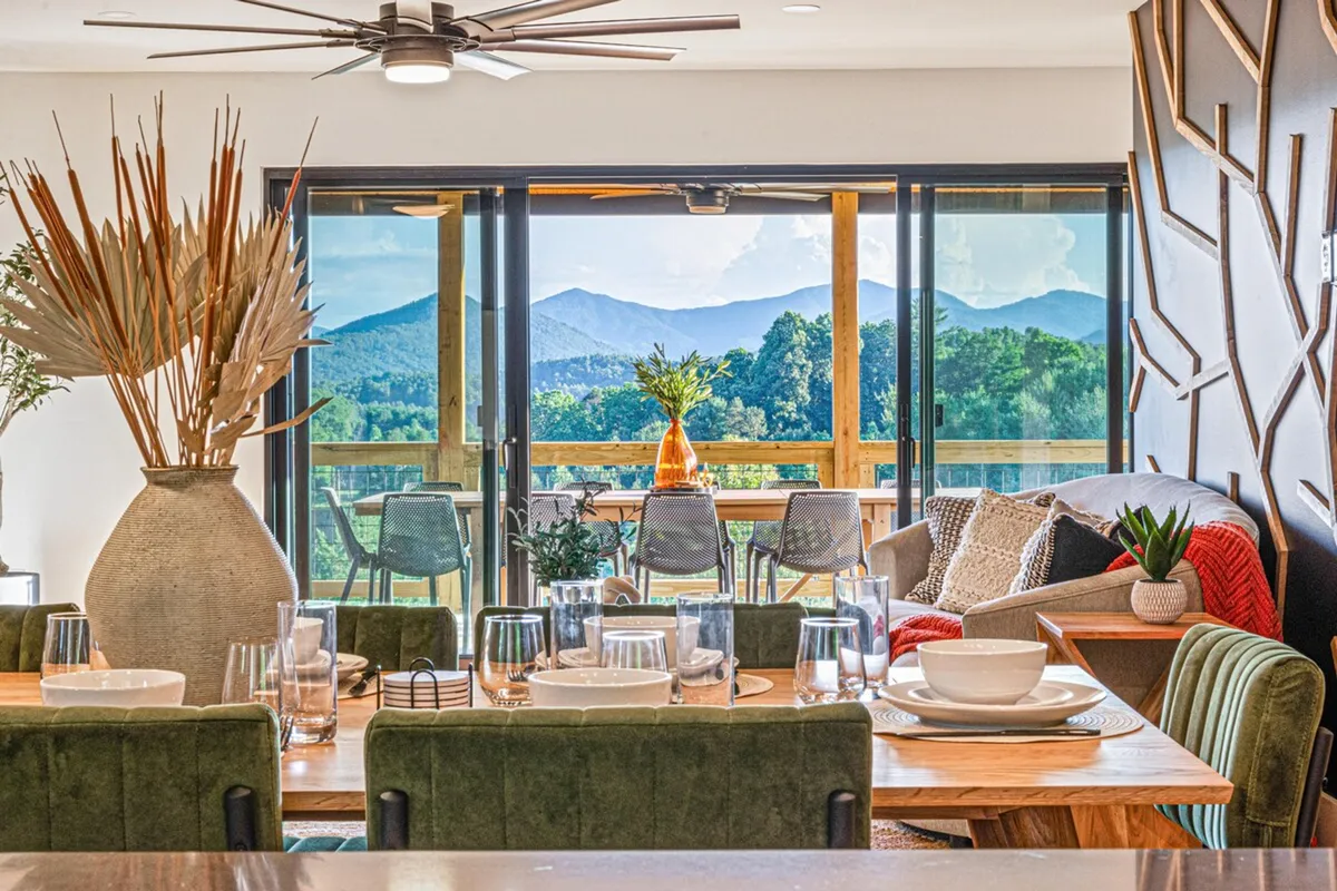 A cozy and inviting dining area with large glass doors that open out to a deck, blending indoor and outdoor living. The dining table is elegantly set, and the comfortable seating area features plush cushions and decorative accents. The space is infused with natural light and offers views of the surrounding landscape.