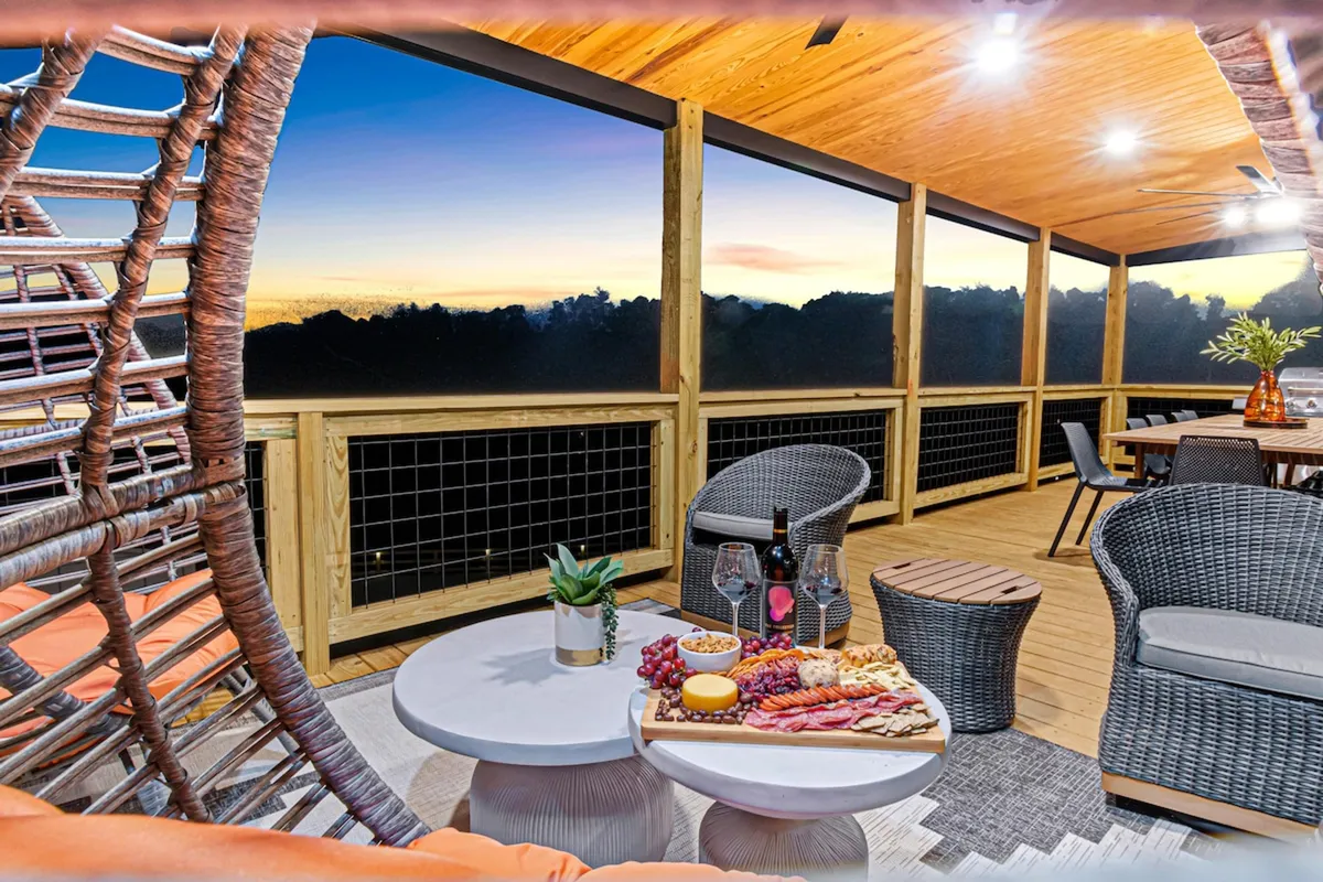 A cozy outdoor dining area featuring a hanging chair, a charcuterie board on a dining table, and space for guests to relax and dine together. The scene is set for intimate or group meals, with sunset views enhancing the atmosphere.