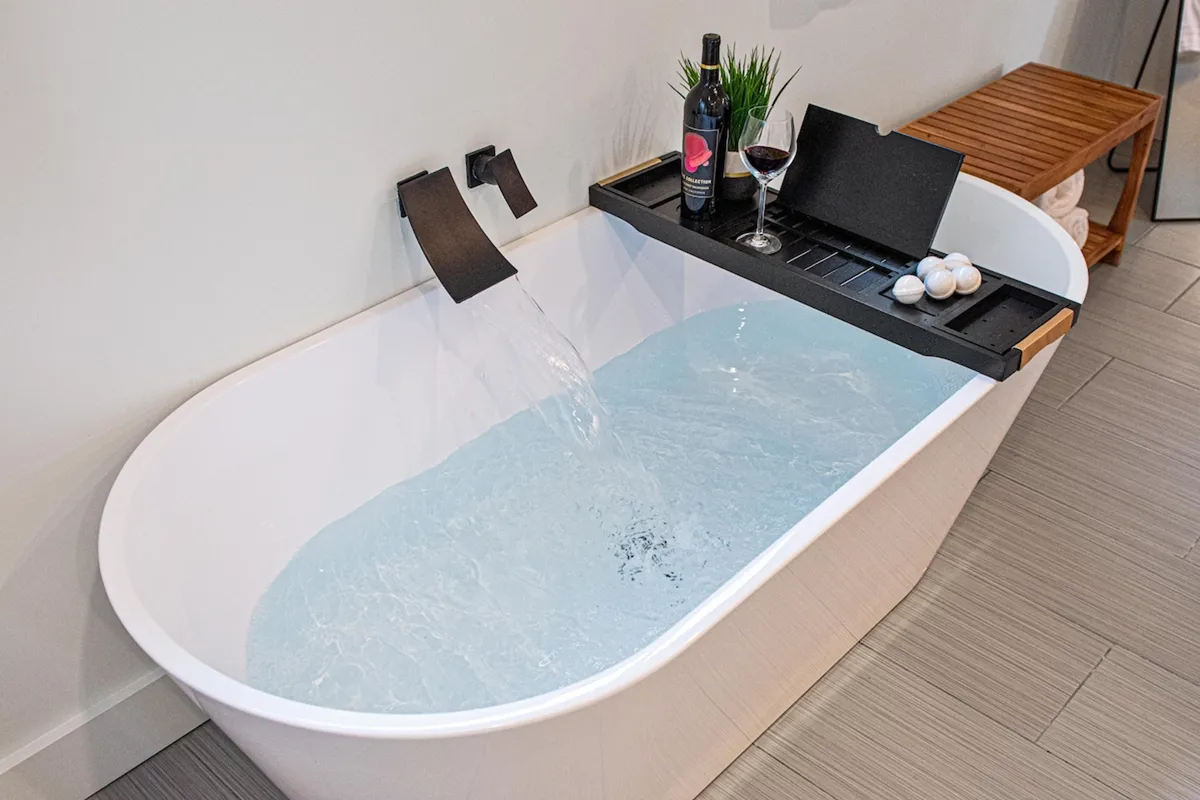 A luxurious bathroom scene featuring a freestanding bathtub with water flowing in, a tray holding a wine glass and elegant décor, and soft, ambient lighting. The scene suggests a space designed for self-care and relaxation.