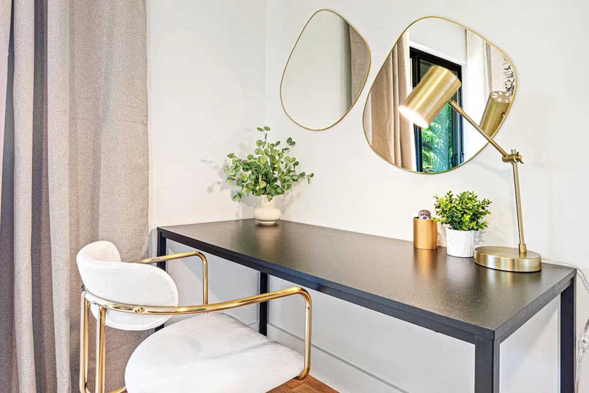 A minimalist desk setup with a golden lamp, mirrors, and potted plants exudes a contemporary, calming atmosphere. The setting invites guests to work, study, or enjoy a quiet moment of reflection.