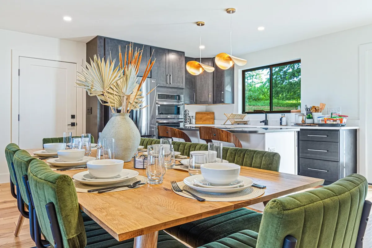 A warm and inviting dining space with a large table set for a meal, seamlessly connected to the kitchen. Natural sunlight streams in through a window, illuminating the decorative plant centerpiece and creating a cozy, communal atmosphere for sharing meals.