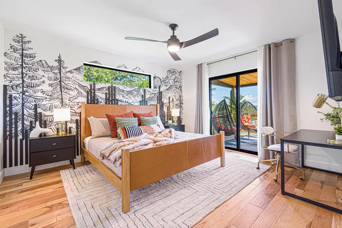 A tranquil bedroom with a wooden bed frame, plush decorative pillows, and large windows revealing serene forest views. Hanging chairs can be seen on an adjacent patio, all bathed in soft, natural light. The décor is warm and earthy, accented by mountain-inspired touches.